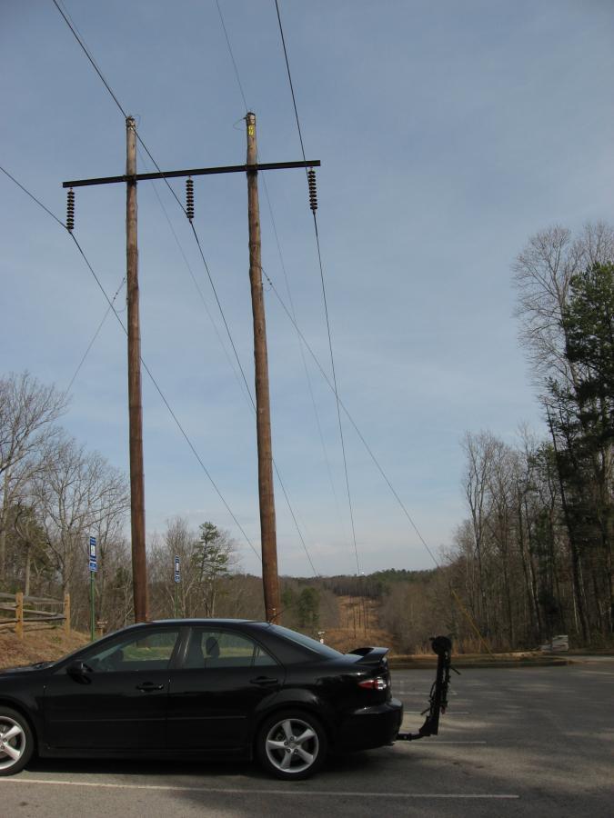 Black car parked by the side of a road with two wooden utility poles and overhead power lines in the background. The scene is set against a clear sky, with sparse trees lining the road, suggesting a rural area. Chicopee Woods mountain bike trail.