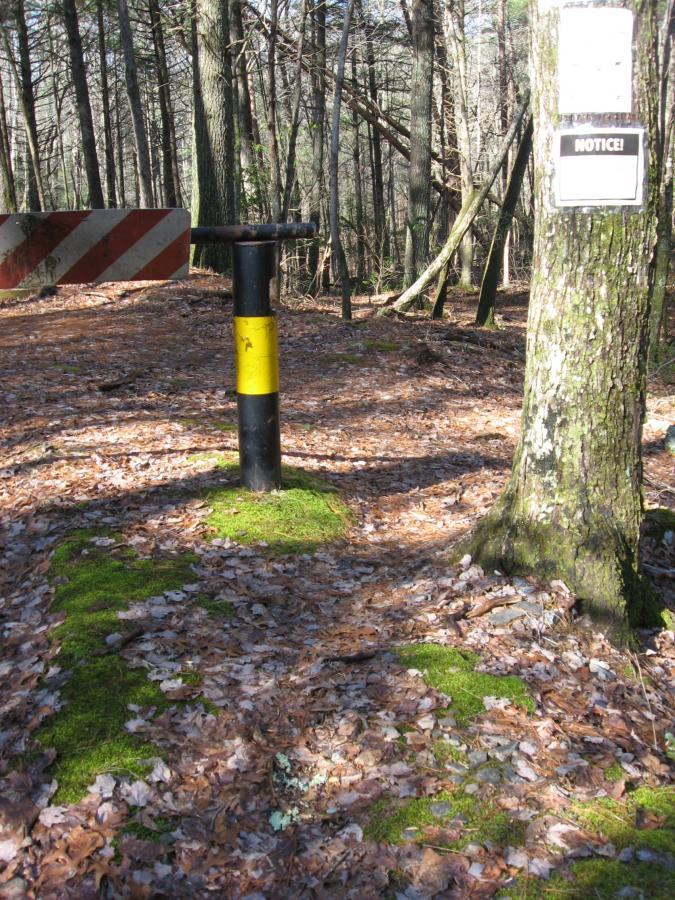 A black and yellow pole in a wooded area, surrounded by fallen leaves and moss. A barrier arm extends from the top of the pole, and a notice sign is attached to a nearby tree. Sunlight filters through the trees, illuminating the peaceful natural setting. Turner Creek Trail mountain bike trail.