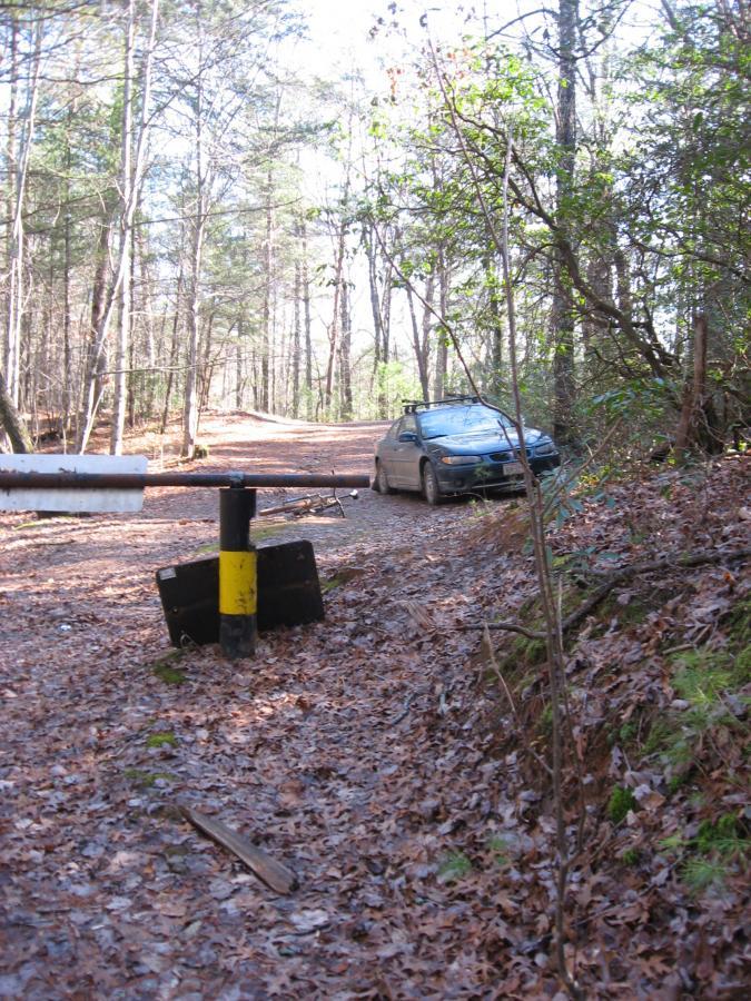 A dirt road surrounded by trees, with a car parked near a barrier made of a yellow and black post. The ground is covered in fallen leaves, and the sunlight filters through the branches overhead. Turner Creek Trail mountain bike trail.