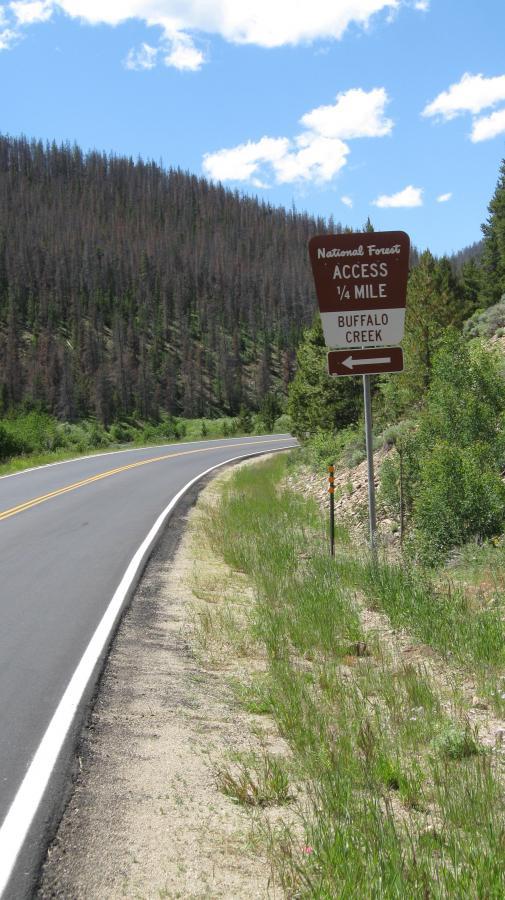 A winding road leading into a national forest, with a sign indicating "National Forest Access ¼ Mile" and directing to "Buffalo Creek." The landscape features green grasses and trees, with a backdrop of hills showing signs of past wildfires. The sky is partly cloudy. Troublesome Creek mountain bike trail.