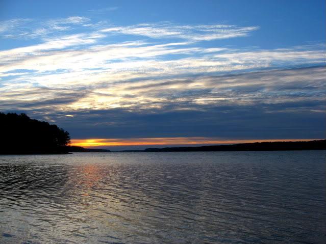 A serene view of a tranquil lake at dusk, with gently rippling water reflecting the colors of the sunset. The sky is adorned with wispy clouds in shades of blue and orange, while a silhouette of trees lines the left side of the image. Hickory Knob State Park mountain bike trail.