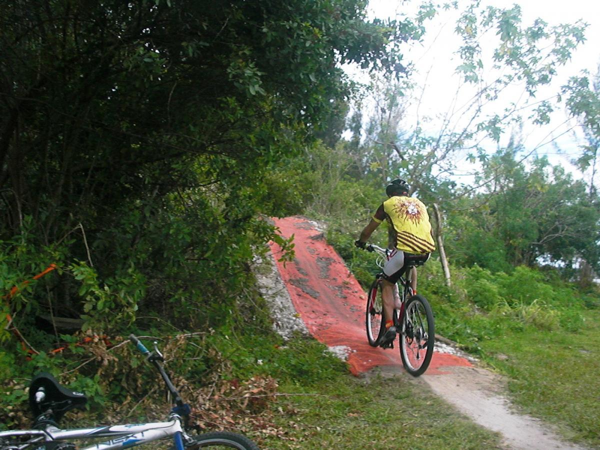 A mountain biker wearing a yellow shirt with a sun design rides up a small red dirt ramp in a wooded area. A second bike is parked nearby, surrounded by greenery and trees in the background. Amelia Earhart Park mountain bike trail.