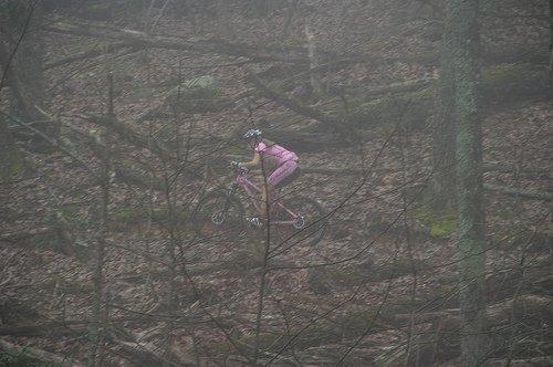 A cyclist in pink attire rides through a foggy forest, navigating over a rugged terrain covered in fallen leaves and branches. The image captures the sense of adventure in a misty, natural setting. Pinhoti Trail mountain bike trail.