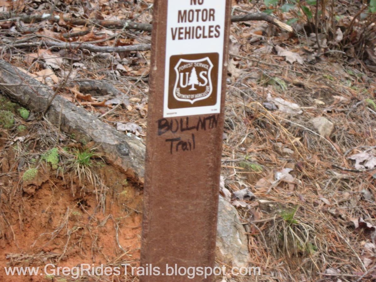 Alt text: A weathered trail sign indicating "BULMNT Trail" with a warning for "No Motor Vehicles" and a U.S. Forest Service logo. The sign is set against a backdrop of forest floor littered with leaves and soil. Bull / Jake Mountain mountain bike trail.