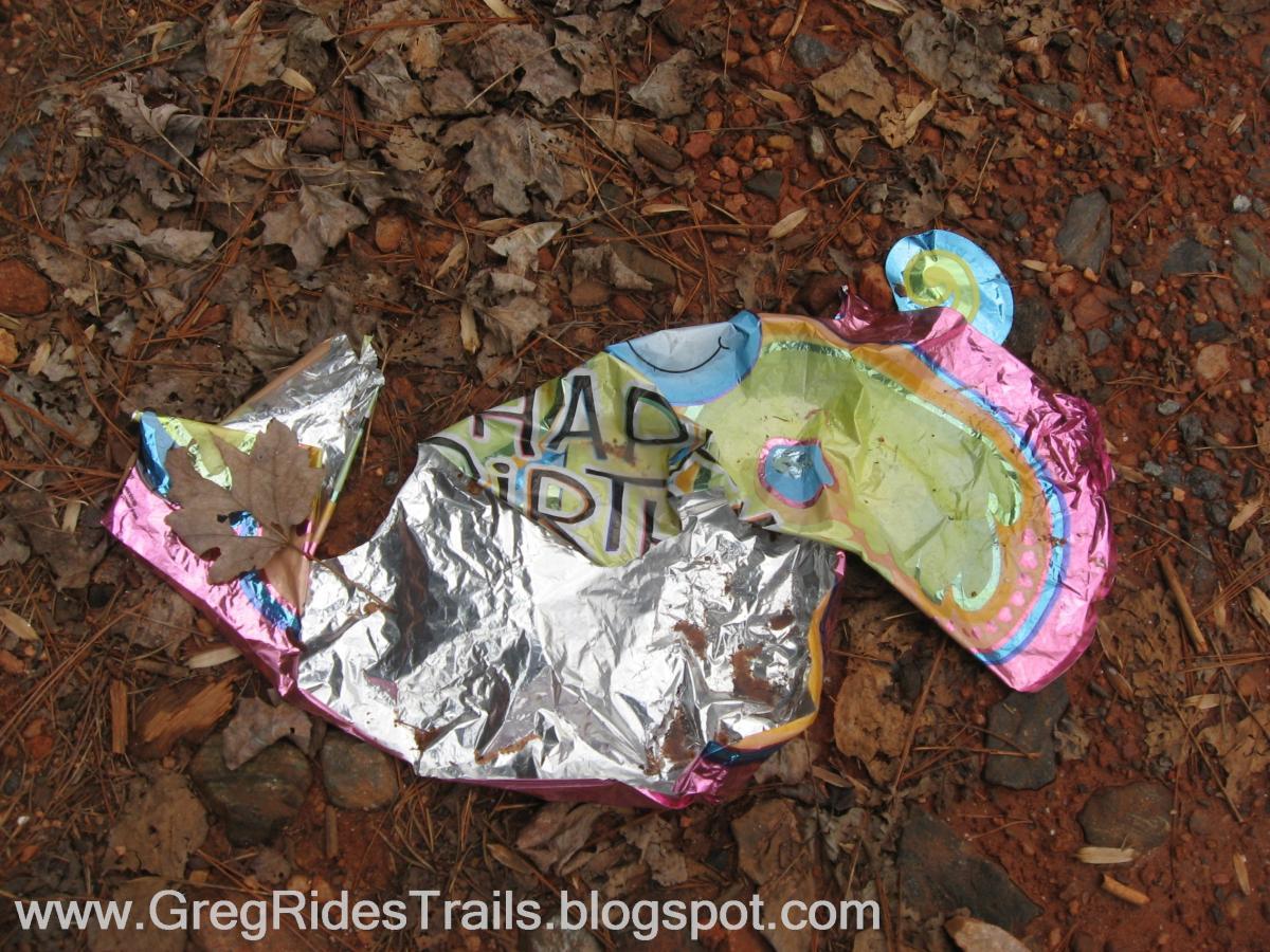 A crumpled, colorful balloon shaped like a fish with "Happy Birthday" written on it, lying on the ground among dried leaves and small rocks. Bull / Jake Mountain mountain bike trail.
