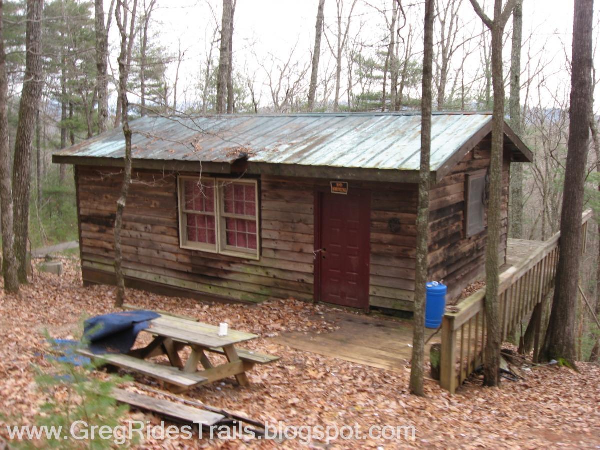 A rustic wooden cabin nestled among trees, with a metal roof and red window frames. The cabin has a porch with a railing and is surrounded by autumn leaves. A wooden picnic table is situated in front, with a blue barrel nearby. Bull / Jake Mountain mountain bike trail.