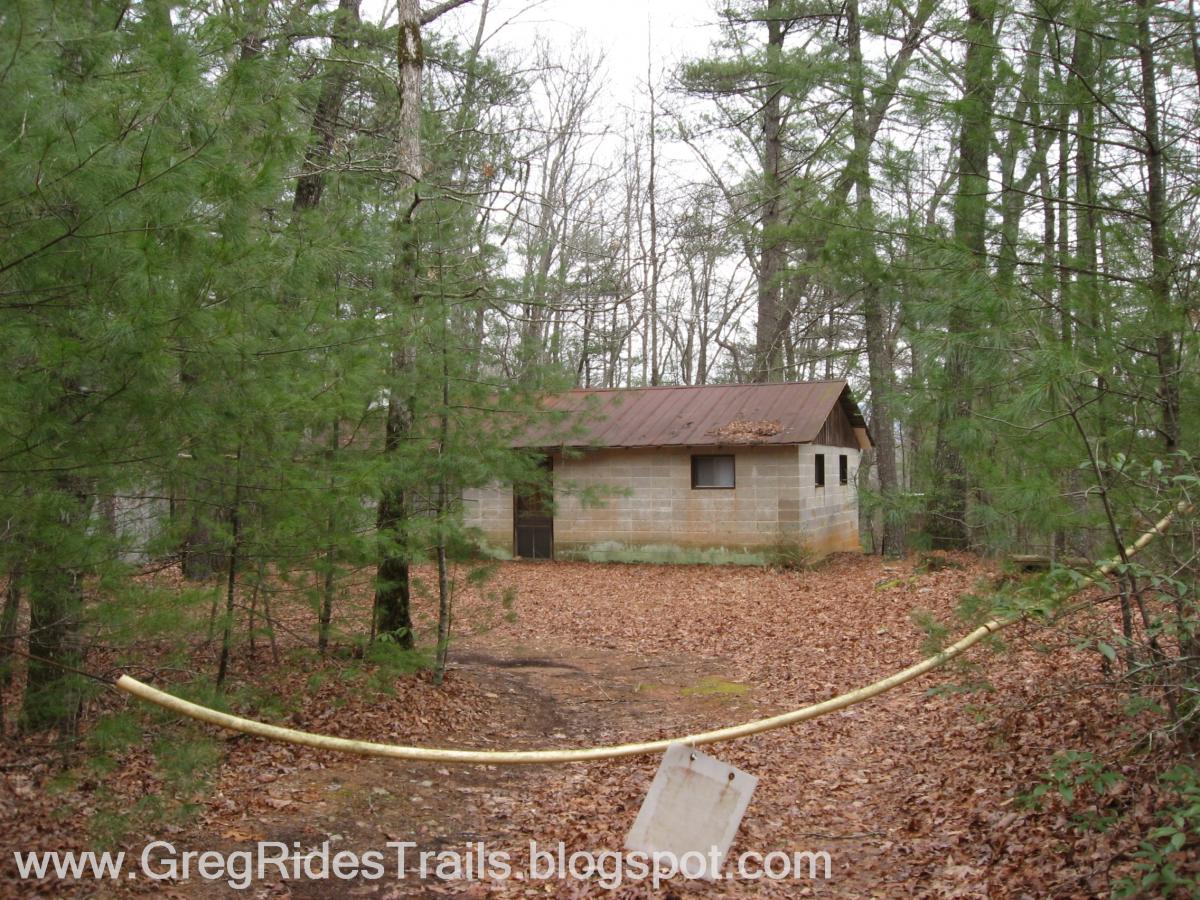 A small, weathered building with a rusty metal roof is nestled in a wooded area, surrounded by tall trees and scattered autumn leaves on the ground. A yellow caution tape stretches across the foreground, indicating restricted access. The building is made of concrete blocks and has a simple window, partially obscured by nearby foliage. Bull / Jake Mountain mountain bike trail.