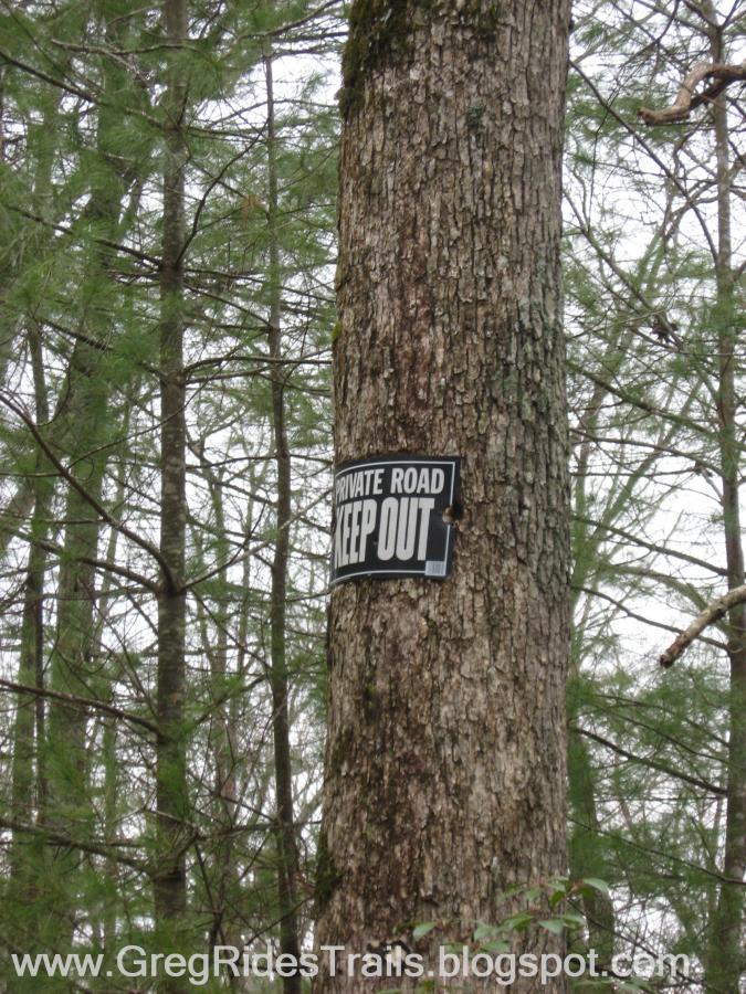 A close-up view of a "Private Road Keep Out" sign affixed to the trunk of a tree, surrounded by dense forest. The sign is black with white lettering, and the background features various green trees and overcast skies. Bull / Jake Mountain mountain bike trail.