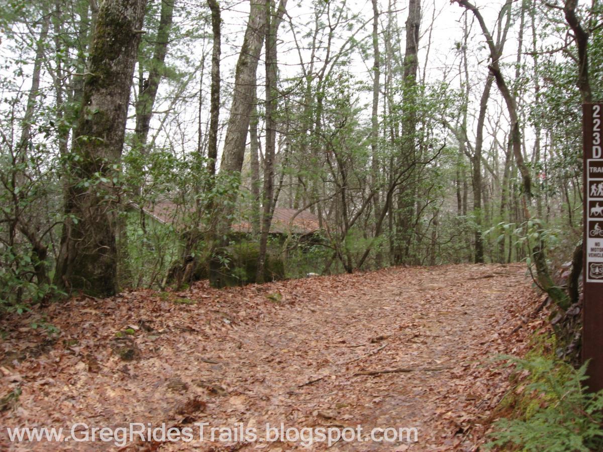 A winding dirt path covered in fallen leaves leads through a wooded area, with tall trees and underbrush lining the sides. In the background, a glimpse of a building with a brown roof is partially obscured by foliage. A trail marker on the right side displays information about the trail and indicates restrictions on motorized vehicles. The atmosphere suggests a serene, natural setting in a forested environment. Bull / Jake Mountain mountain bike trail.