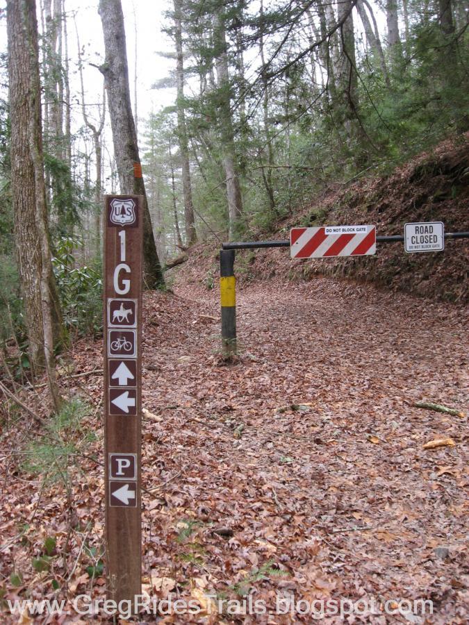 A forest trail intersection sign showing direction indicators for horseback riding, biking, and parking, alongside a closed road gate with warning signs. The ground is covered with dried leaves, and surrounding trees create a wooded environment. Bull / Jake Mountain mountain bike trail.