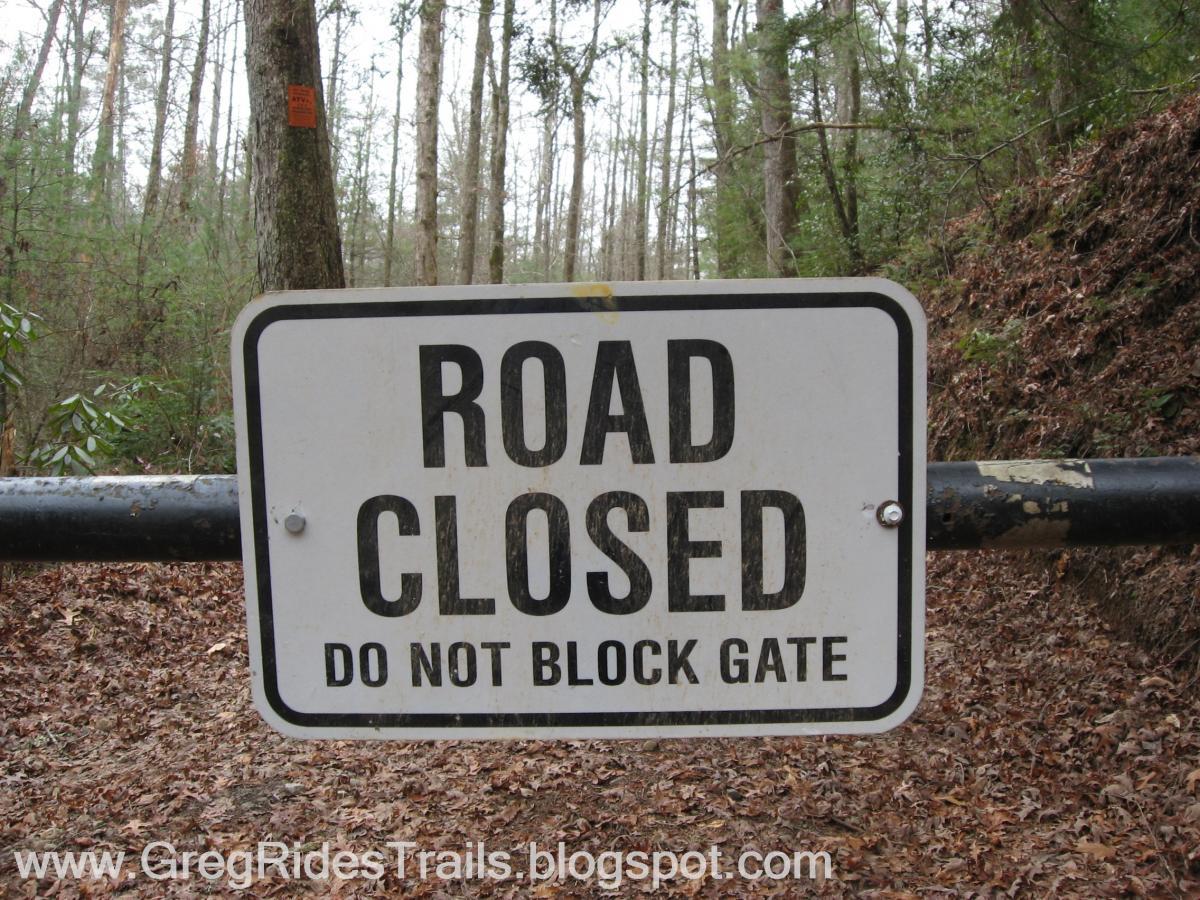 A road closed sign on a metal gate in a forested area, stating "ROAD CLOSED DO NOT BLOCK GATE." Trees and fallen leaves are visible in the background. Bull / Jake Mountain mountain bike trail.