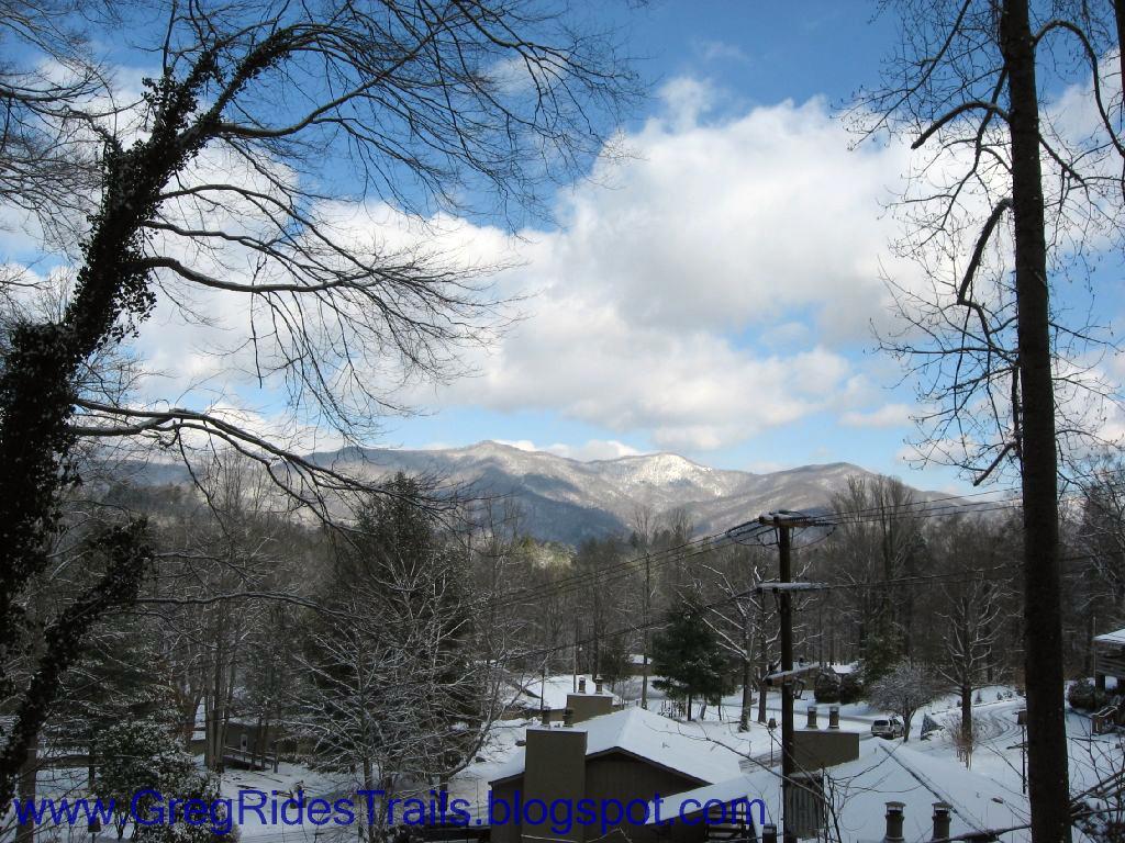 A winter landscape featuring snow-covered rooftops and bare trees in the foreground, with distant mountains under a partly cloudy blue sky. The scene conveys a peaceful, serene atmosphere typical of a snowy day in the mountains. Fontana Village mountain bike trail.