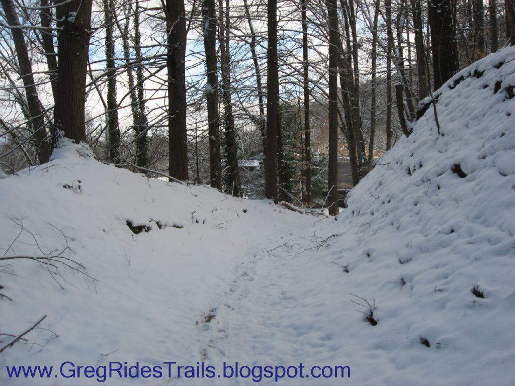 A snowy forest trail with tall trees lining both sides, creating a peaceful, serene atmosphere. The ground is covered in a blanket of snow, and there are subtle footprints leading down the path, suggesting recent activity. The light filters softly through the branches, adding to the tranquil winter scene. Fontana Village mountain bike trail.