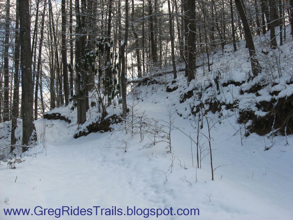 A snowy forest path winding through trees, with a rocky incline on one side and a blanket of fresh snow covering the ground. The scene captures the serene beauty of nature in winter, with leafless trees and a clear, bright sky above. Fontana Village mountain bike trail.