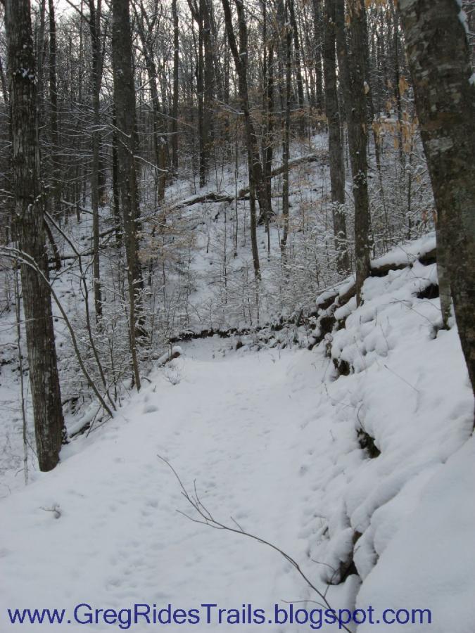 A snow-covered hiking trail winding through a wooded area with bare trees, showcasing a serene winter landscape. The path leads into the distance, flanked by snow-dusted ground and rocky edges. Fontana Village mountain bike trail.