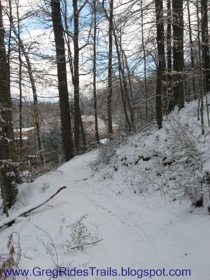 A snow-covered trail winding through a forest of bare trees, with patches of blue sky peeking through the clouds. The scene captures a peaceful winter landscape, inviting outdoor exploration. Fontana Village mountain bike trail.