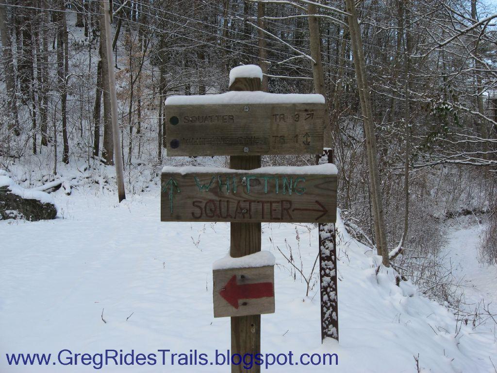 A wooden trail sign in a snowy forest, indicating directions for 'Squatter Trail' and 'Whipping Squatter' with distance markers and arrows. The surrounding trees are bare, showcasing a winter landscape. Fontana Village mountain bike trail.