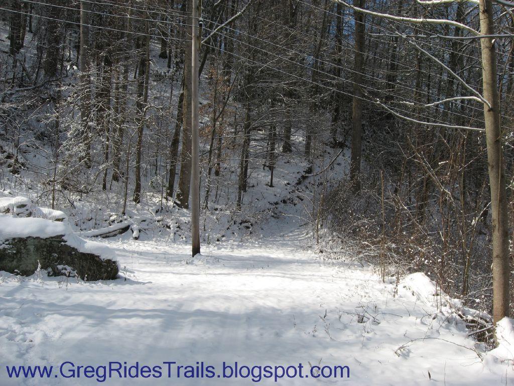 A snow-covered trail winding through a forest, with trees lining both sides and a clear blue sky visible above. The ground is blanketed with fresh snow, and a utility pole is spotted along the path. Fontana Village mountain bike trail.