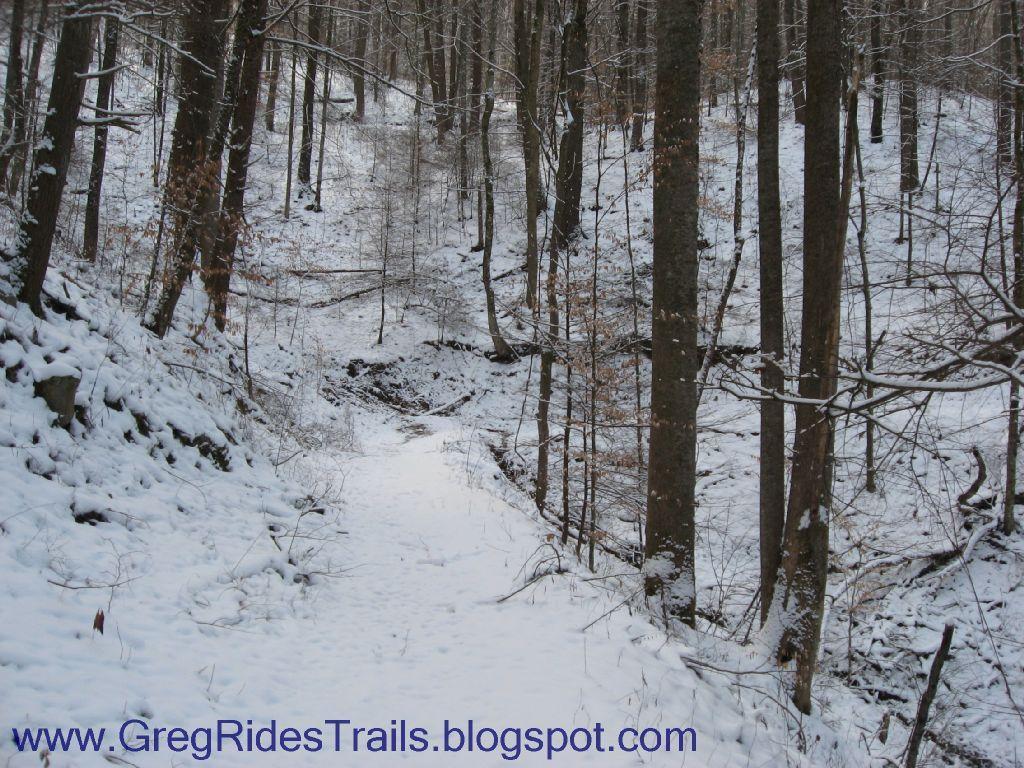 A snow-covered trail winding through a forest, with tall trees on either side and a gentle slope leading down into a snowy clearing. The ground is blanketed in fresh snow, creating a serene winter landscape. Fontana Village mountain bike trail.