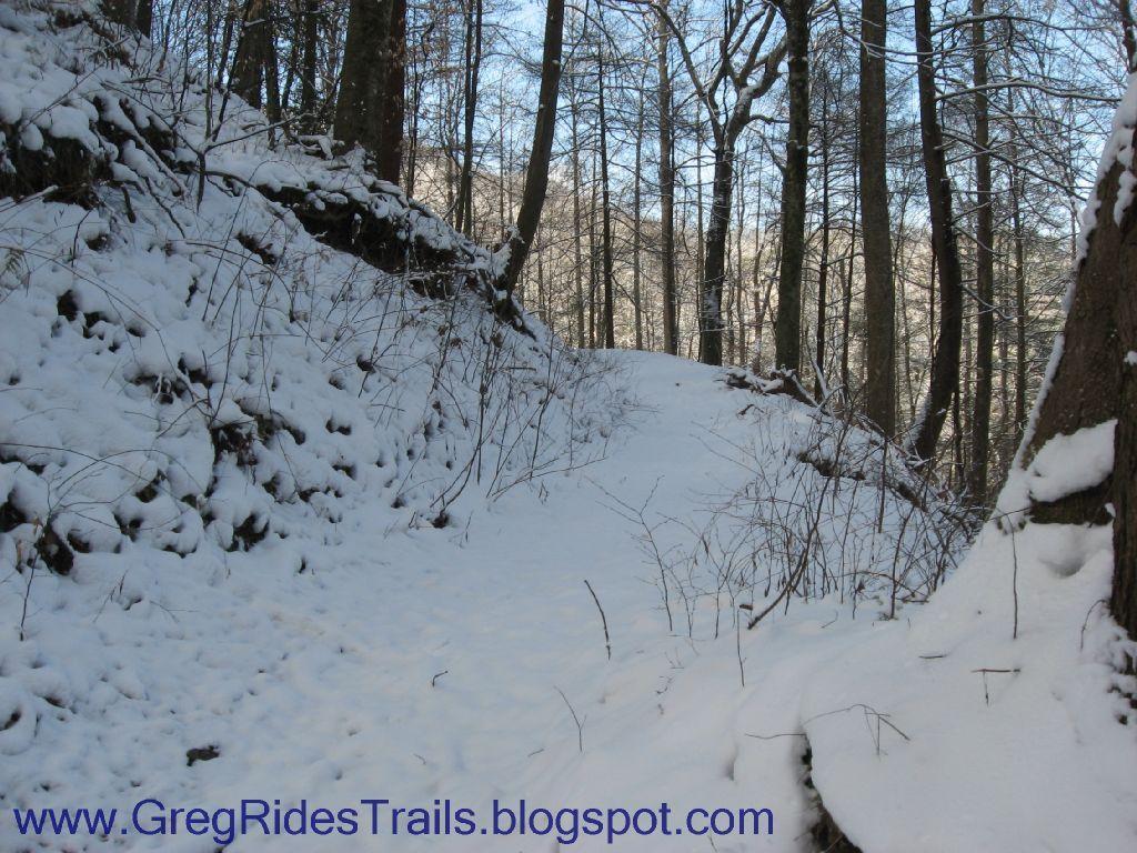 A snowy trail winding through a forest, with trees lining both sides and a blanket of fresh snow covering the ground. The scene evokes a peaceful winter atmosphere, highlighting the natural beauty of the landscape. Fontana Village mountain bike trail.