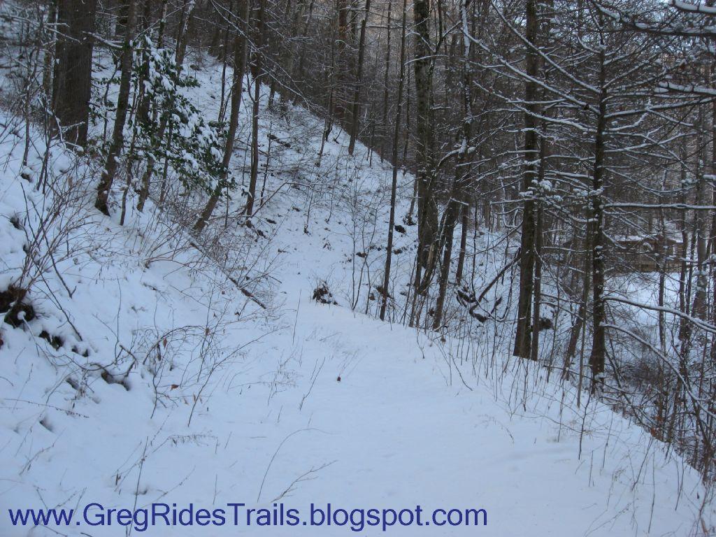 A snowy trail winding through a forest of bare trees and evergreen plants, with a snow-covered ground and a serene, tranquil atmosphere. Fontana Village mountain bike trail.