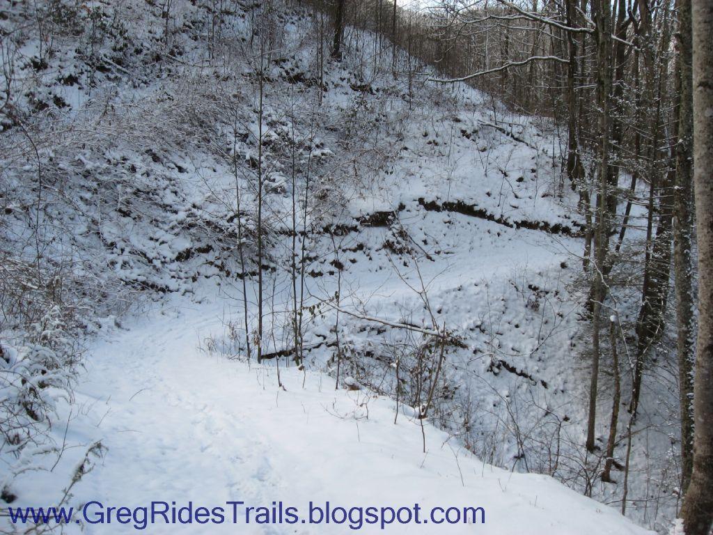 A snowy path winds through a forested area, flanked by trees. The ground is covered in a blanket of snow, and there are hints of vegetation poking through. The landscape features a hillside with exposed earth and a gentle slope leading into a nearby valley. Fontana Village mountain bike trail.