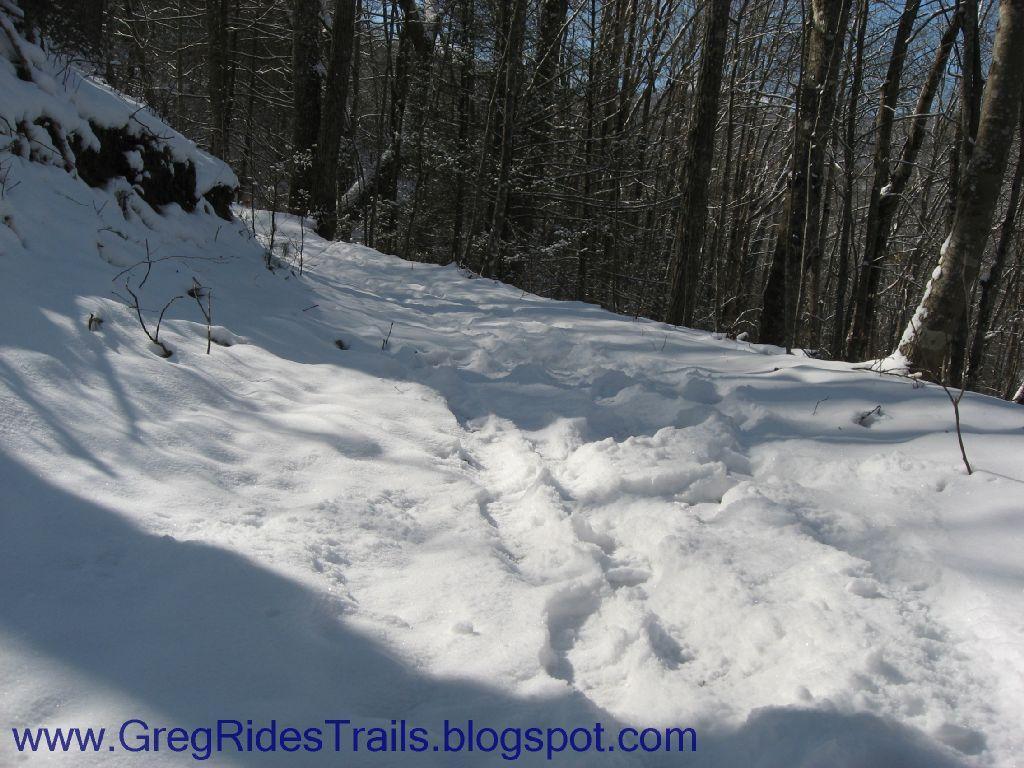 A snow-covered trail winding through a wooded area, with footprints visible in the fresh snow. The trees are bare, indicating winter, and sunlight filters through the branches, casting soft shadows on the ground. Fontana Village mountain bike trail.