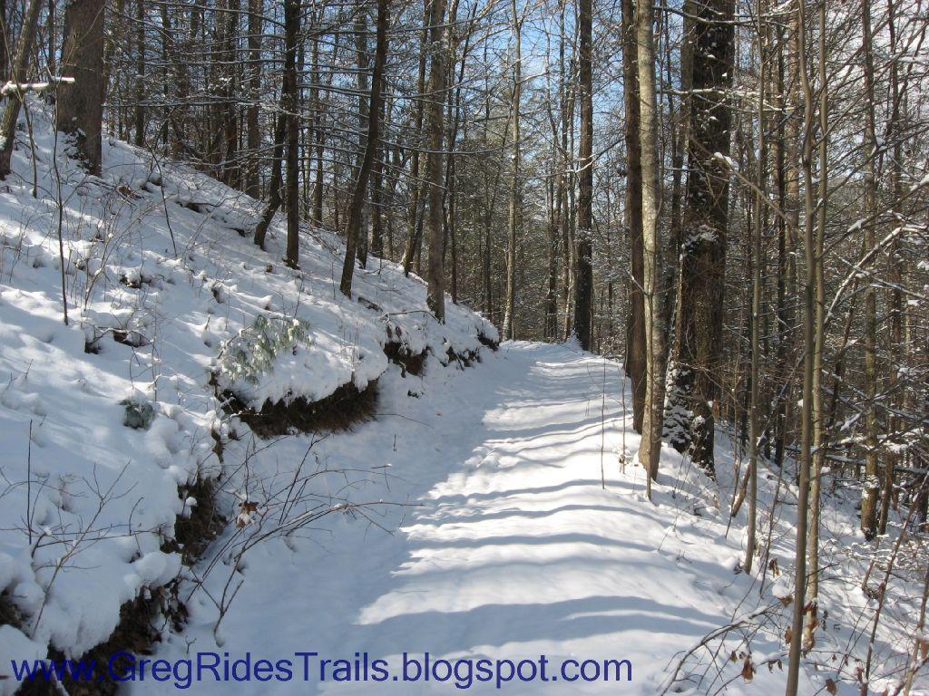 A snowy trail winding through a forest of bare trees. The ground is covered in a thick layer of fresh snow, with sunlight filtering through the branches, creating shadows on the path. Fontana Village mountain bike trail.