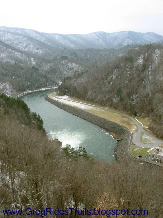 A scenic view of a winding river surrounded by mountains, with a clear sky and patches of snow on the peaks. The riverbank features a grassy area and a small parking lot, set among bare trees in the foreground. Fontana Village mountain bike trail.