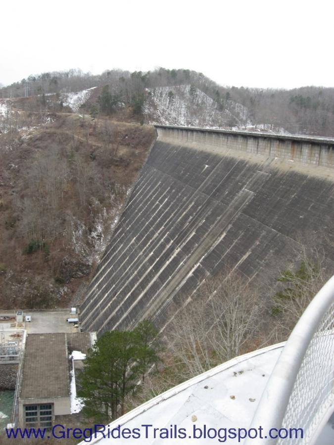 Alt text: A steep concrete dam rises from a valley, surrounded by bare trees and rolling hills under a cloudy sky. A portion of the dam's base is visible, along with a building situated at the base. The scene conveys a sense of scale and engineering amidst a natural landscape. Fontana Village mountain bike trail.