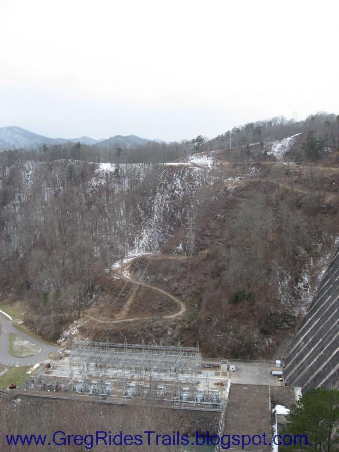 A panoramic view of a mountainous landscape featuring a dam infrastructure in the foreground. In the lower part of the image, there are electrical equipment structures, while the background showcases a hillside with sparse trees and a winding dirt road leading up into the mountains. The scene has a cloudy sky, suggesting a cool, overcast day, with patches of snow visible on the slopes. Fontana Village mountain bike trail.