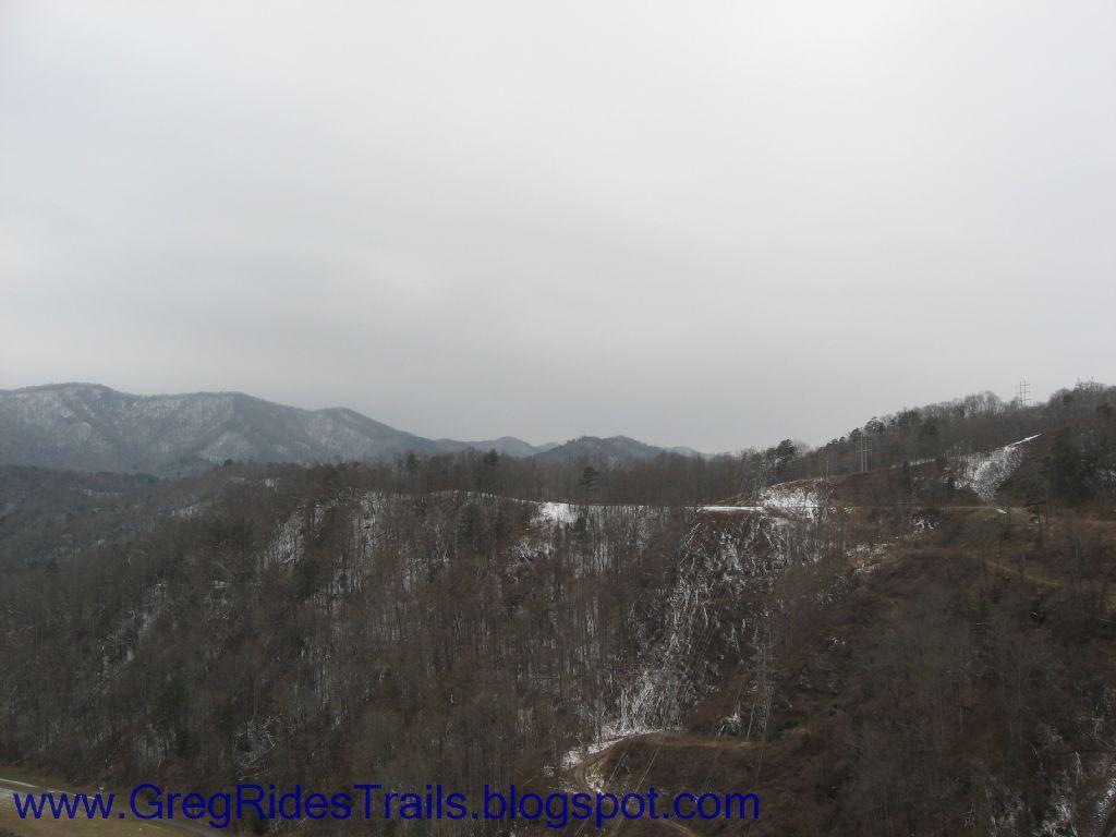 A panoramic view of misty mountains under a cloudy sky, featuring snow-dusted peaks and a forested landscape with bare trees. The scene conveys a tranquil winter atmosphere, showcasing the rugged beauty of nature. Fontana Village mountain bike trail.
