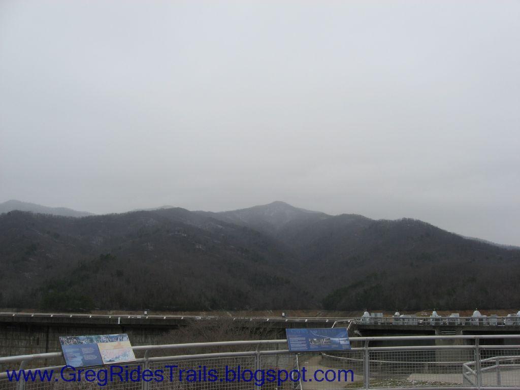 A scenic view of a mountain landscape under a cloudy sky, with a dam visible in the foreground. There are informational signs in the lower left corner detailing the area. The mountains are partially obscured by clouds, creating a misty atmosphere. Fontana Village mountain bike trail.