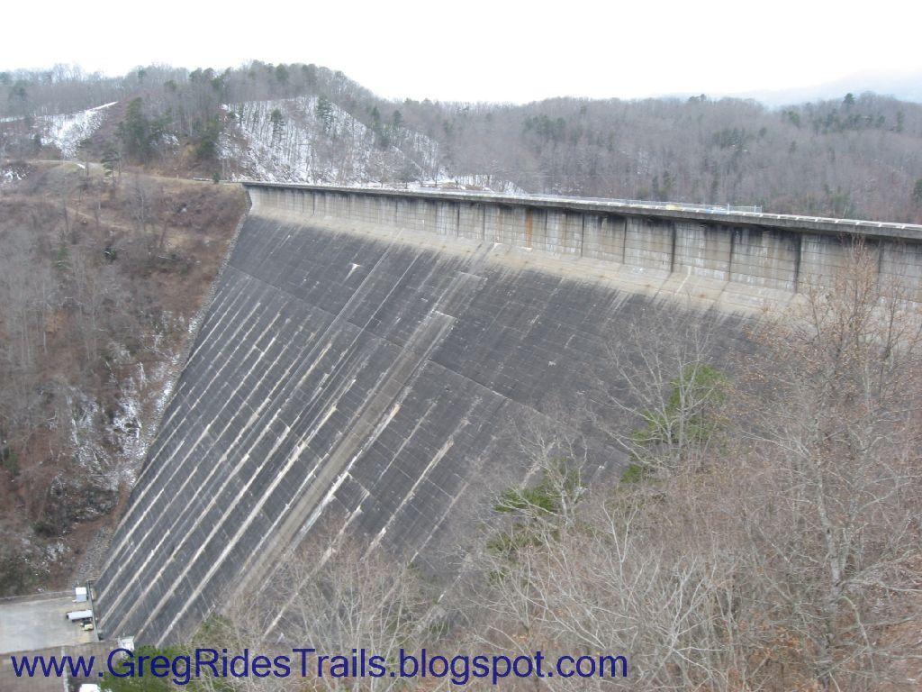 A large concrete dam rises steeply against a backdrop of wooded hills. The dam's surface is marked by weathering and wear, and a narrow roadway runs along the top. In the foreground, sparse vegetation is visible, indicating a colder season, with some areas covered in light snow. The overall scene conveys a sense of scale and the engineered landscape. Fontana Village mountain bike trail.