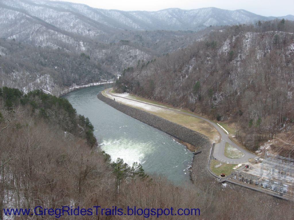 A scenic view of a river winding through a mountainous landscape, with a dam and a road visible along the riverbank. Surrounding the area are trees, some of which are dusted with snow, and distant mountains shrouded in mist. Fontana Village mountain bike trail.