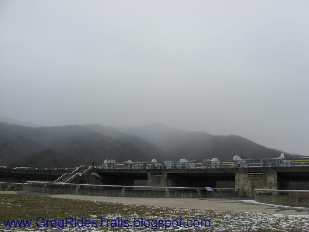 A view of a dam under overcast skies, with misty mountains in the background. The dam features concrete structures and gates, while the foreground includes grassy areas that appear to be lightly dusted with snow. Fontana Village mountain bike trail.