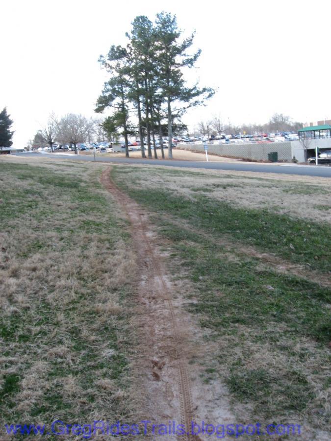 A dirt path winding through a grassy area, leading towards a cluster of trees in the background. A parking lot and a building are visible further back, indicating an urban or suburban setting. Gainesville College mountain bike trail.