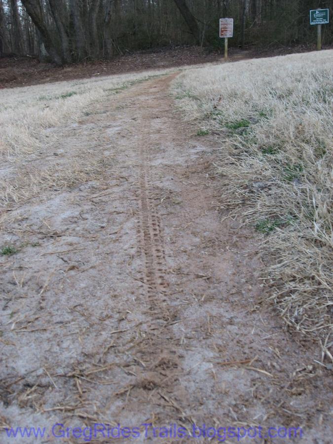 A dirt path leading into a wooded area, flanked by dry grass on either side. Tire tracks are visible in the center of the path, suggesting recent activity. Signs indicating information about the area can be seen nearby. Gainesville College mountain bike trail.