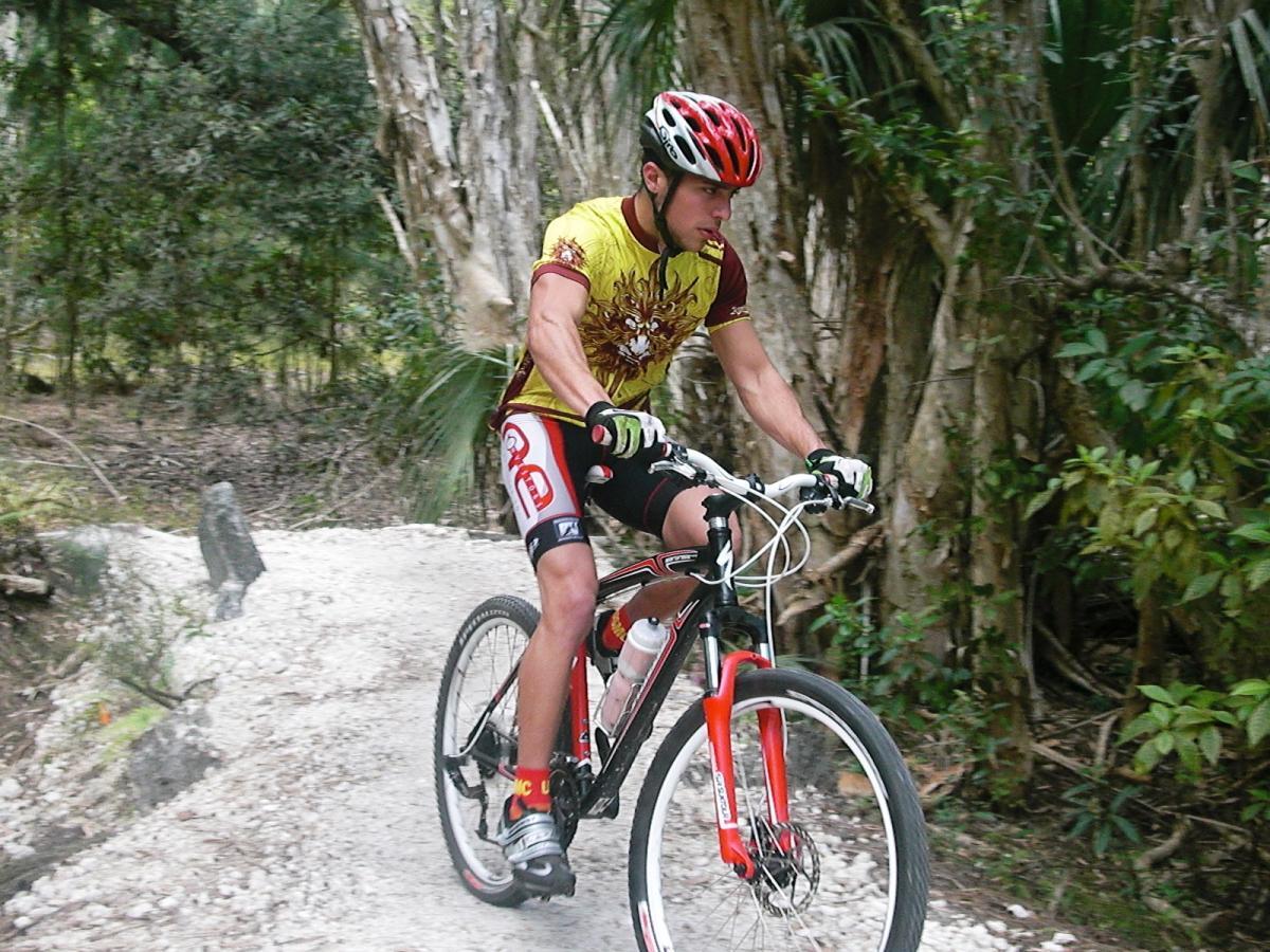 A cyclist wearing a yellow and red jersey rides a mountain bike on a gravel path surrounded by lush greenery and trees. The cyclist is focused and in motion, showcasing an active outdoor lifestyle. Amelia Earhart Park mountain bike trail.