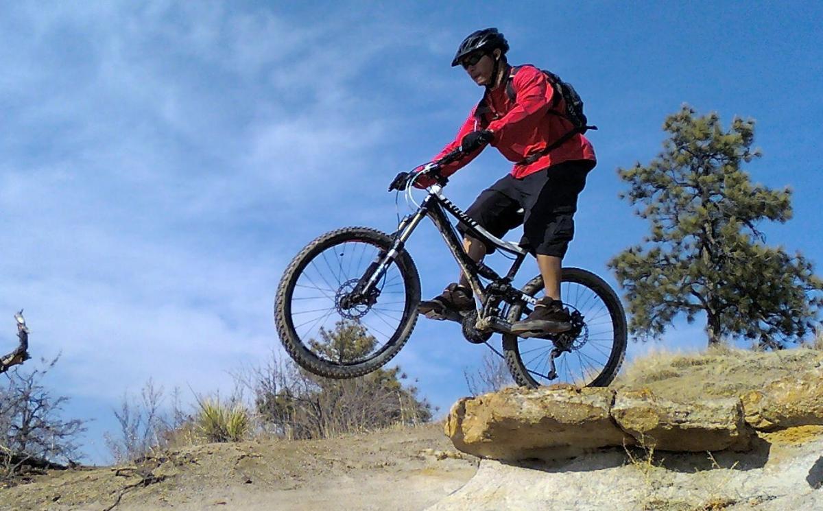 A mountain biker in a red jacket jumps off a rocky ledge while riding a black bicycle, with a blue sky and sparse trees in the background. Palmer Park mountain bike trail.