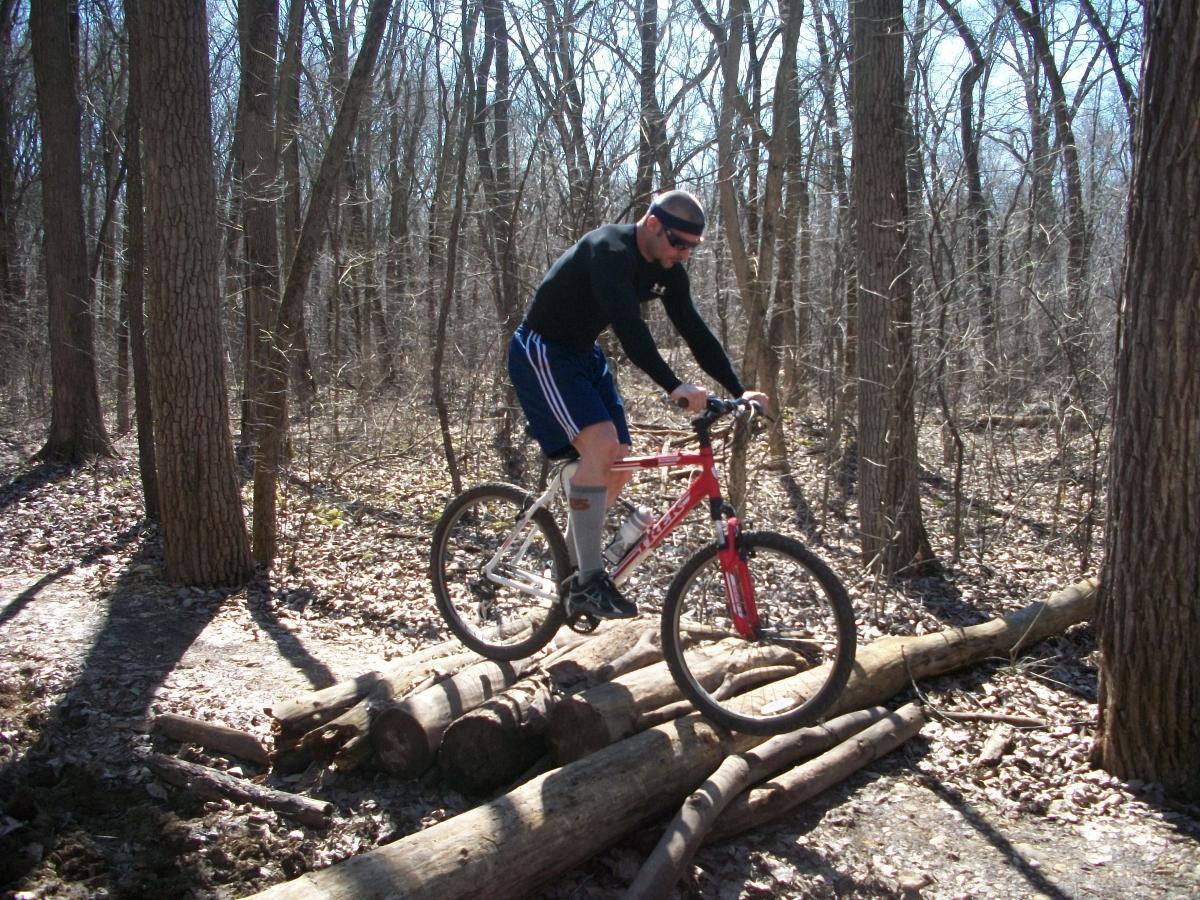 A mountain biker navigating over fallen logs in a forested area during daytime. The rider is wearing a black long-sleeve shirt, blue shorts, and knee-high socks, with a headband and sunglasses. Trees with bare branches are visible in the background, indicating early spring or late winter. Saw Wee Kee Park mountain bike trail.