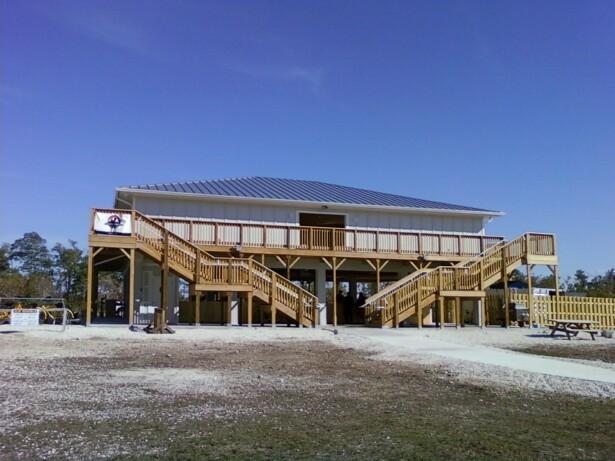 A large, two-story building with a metal roof, featuring wooden decks and staircases on both sides. The structure is set against a clear blue sky and is surrounded by a gravel area. There is a sign visible on the front of the building. Oleta River State Park mountain bike trail.