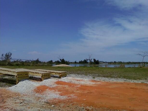 A wide view of an open landscape featuring wooden structures arranged on a grassy area, with piles of gravel and orange soil in the foreground. In the background, a calm body of water and trees are visible under a clear blue sky with scattered clouds. Quiet Waters Park mountain bike trail.