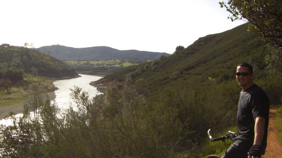 A man in a black t-shirt and sunglasses poses with a bicycle on a dirt path overlooking a serene river surrounded by lush green hills. The sun shines in a clear sky, highlighting the natural landscape. Sweetwater Trail mountain bike trail.