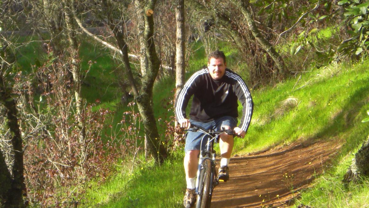 A man biking on a dirt trail surrounded by greenery and trees on a sunny day. He is wearing a black long-sleeve shirt with white stripes and blue shorts. Sweetwater Trail mountain bike trail.