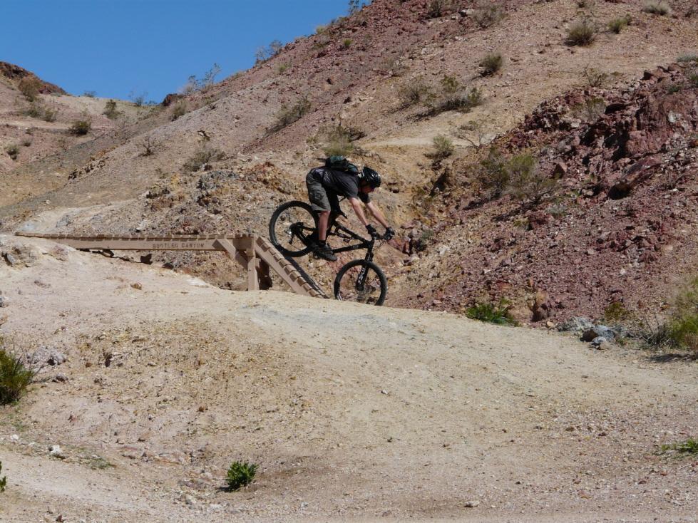 A mountain biker performing a jump over a wooden ramp on a dirt trail, surrounded by rocky terrain and sparse vegetation under a clear blue sky. Bootleg Canyon mountain bike trail.
