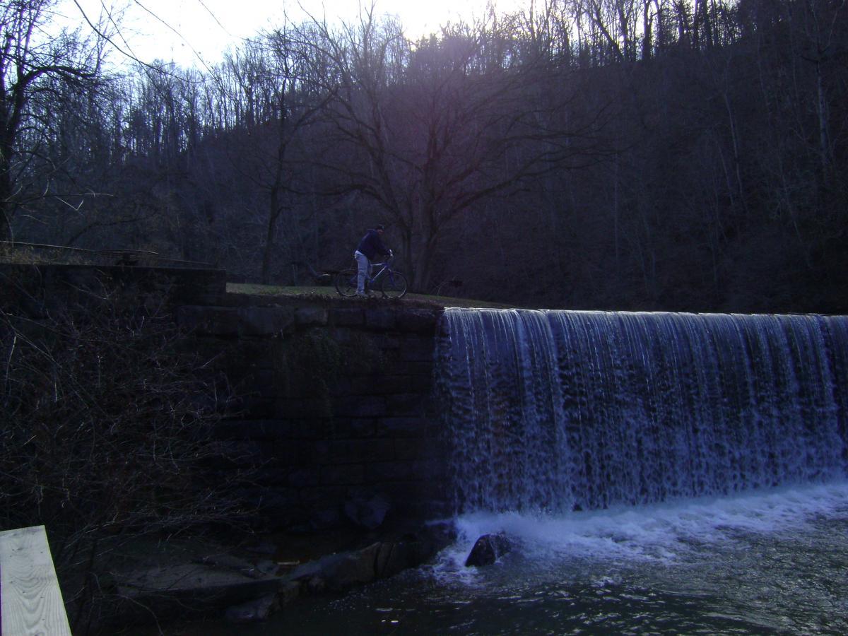 A person stands with a bicycle near a stone wall beside a waterfall, surrounded by bare trees and a hilly landscape. The scene is set on a cloudy day, with the water cascading over the edge of the wall into a river below. Black Water Creek mountain bike trail.