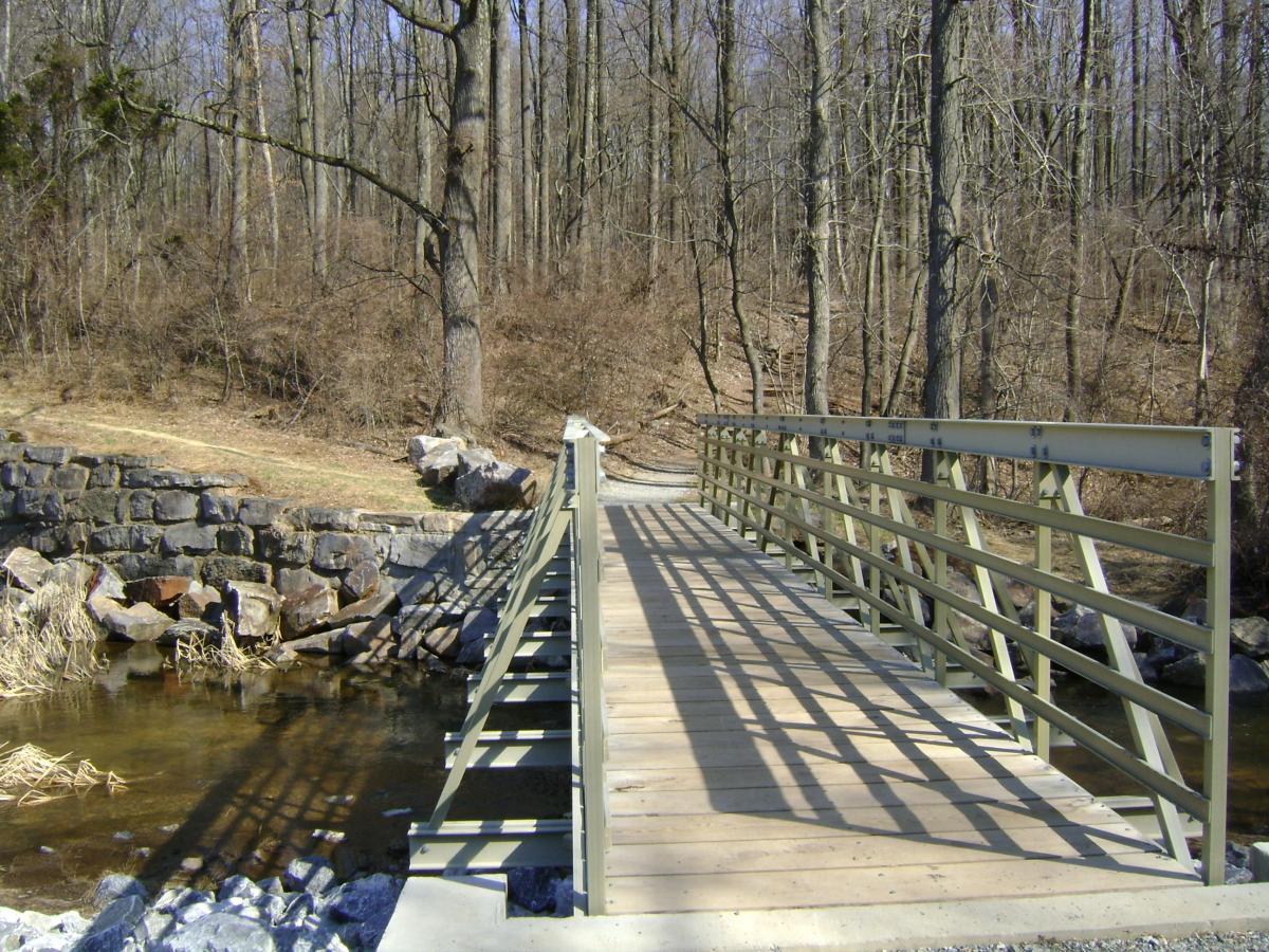 A metal bridge crossing a small stream, surrounded by bare trees and rocky banks, with a pathway leading into the forest. Sunlight casts shadows on the bridge, highlighting its structure against the serene natural backdrop. French Creek mountain bike trail.