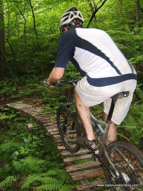 A cyclist riding a mountain bike on a narrow wooden boardwalk trail, surrounded by lush green foliage in a forest setting. The rider is wearing a helmet and sports attire, with mud splattered on the bike and clothing, indicating recent activity on a rugged path. Liberty Mountain mountain bike trail.
