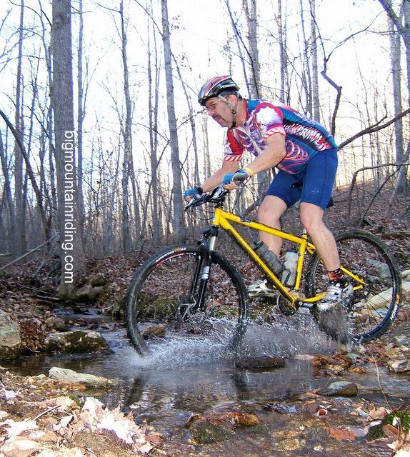A cyclist in a colorful jersey rides a yellow mountain bike through a shallow stream in a forested area, splashing water as he navigates over rocks and fallen leaves. Bare trees can be seen in the background, indicating early spring or fall. Liberty Mountain mountain bike trail.